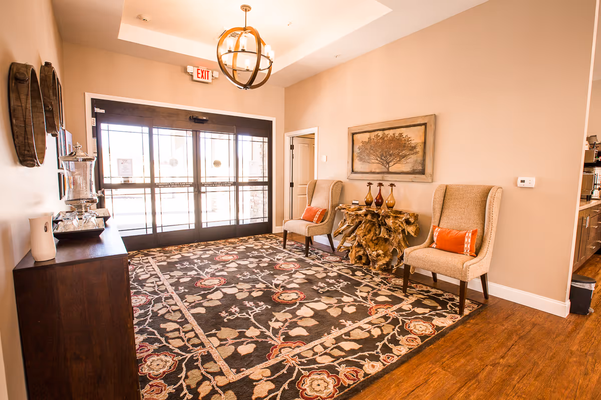 A warmly lit entrance area with double glass doors, a large floral patterned rug, two beige upholstered chairs with orange accent pillows, a unique wooden console table with decorative vases, and a framed tree artwork on the wall.