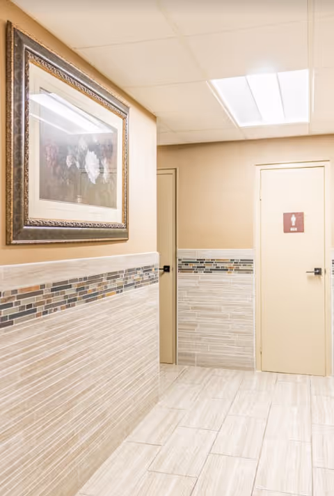 Interior hallway with beige tiled walls and floor, a framed floral painting on the left wall, and two closed doors at the end of the hallway. One door has a sign indicating it is a men's restroom.