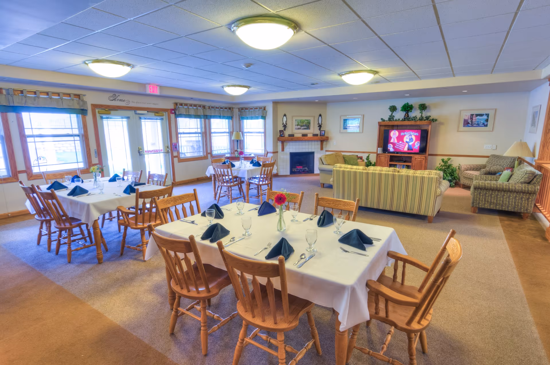 A bright and spacious dining area with several wooden tables covered with white tablecloths, each set with glassware, silverware, and folded dark napkins. The room has large windows with curtains, a fireplace, and a cozy seating area with sofas and a television in the background. The walls are decorated with framed pictures and plants.