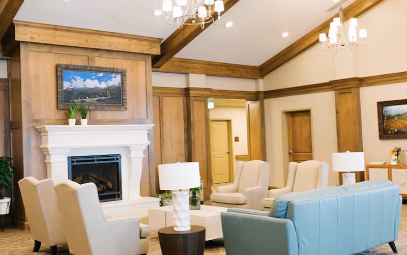 Spacious furnished common living room with sofas, armchairs, coffee tables, a fireplace, and wood-paneled walls.