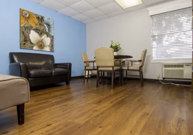 A room with wooden flooring featuring a black leather loveseat on the left, a round table with four beige upholstered chairs in the center, a blue accent wall with a large floral painting, and a window with blinds and an air conditioning unit on the right.