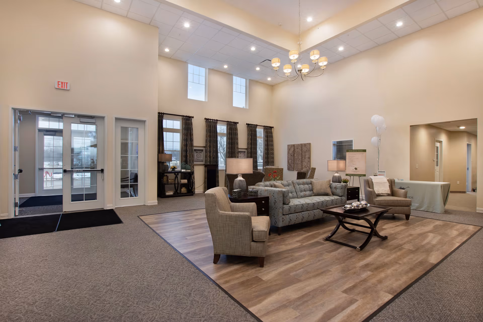 A spacious and well-lit living room area with high ceilings and large windows. The room features a seating arrangement with a patterned sofa, two armchairs, a coffee table, and side tables with lamps. The floor has a combination of carpet and wood-like flooring. There is an exit door with glass panels on the left side and a table with balloons and a sign on the right side near a hallway.