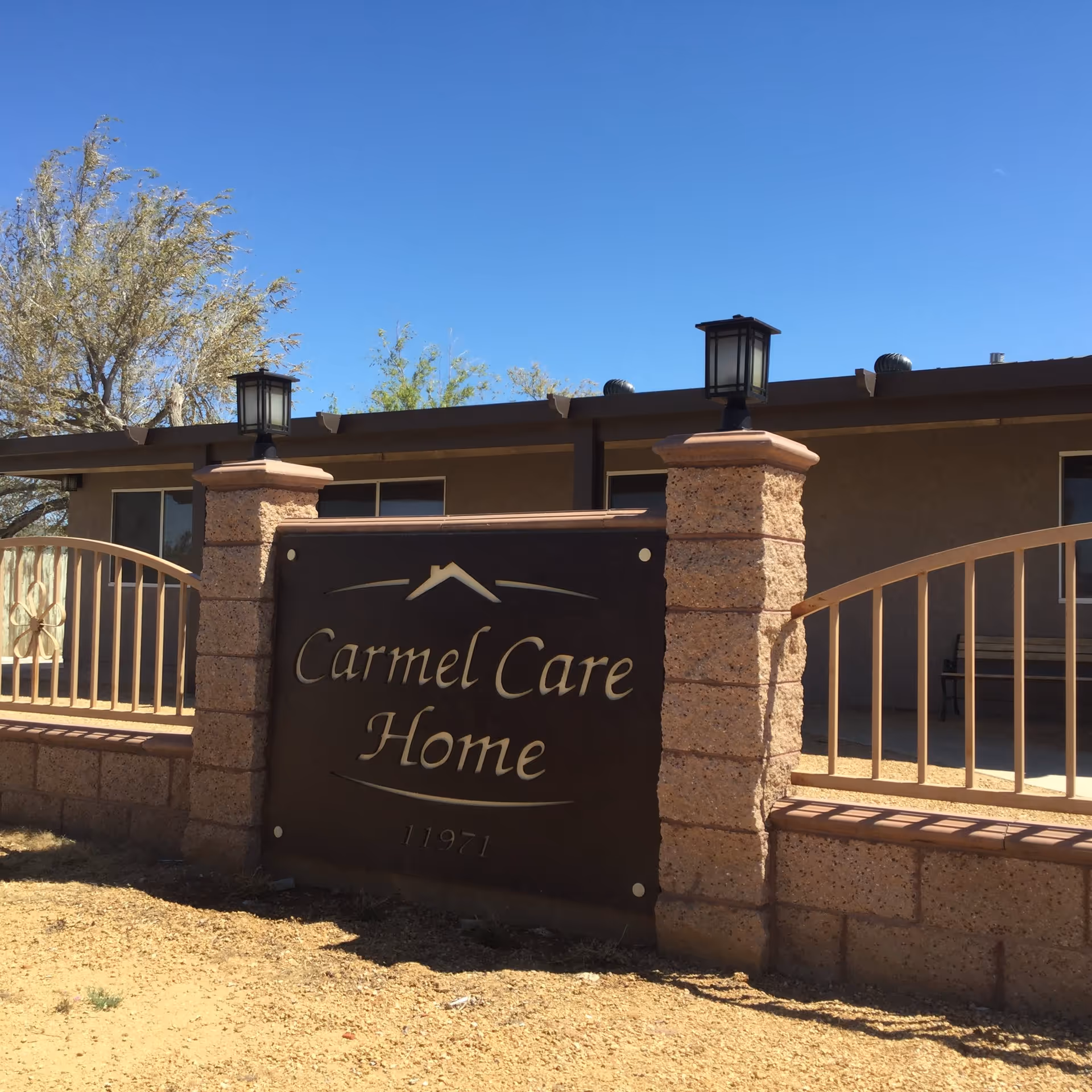 Outdoor view of the entrance sign for Carmel Care Home, featuring a brown sign mounted on a stone and metal fence with two lantern-style lights on top of stone pillars, under a clear blue sky.