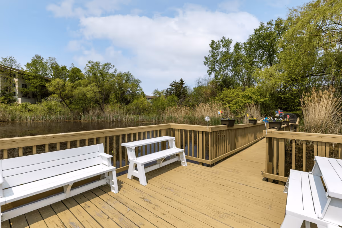 A wooden deck with white benches overlooking a pond surrounded by tall grasses and trees under a partly cloudy sky.
