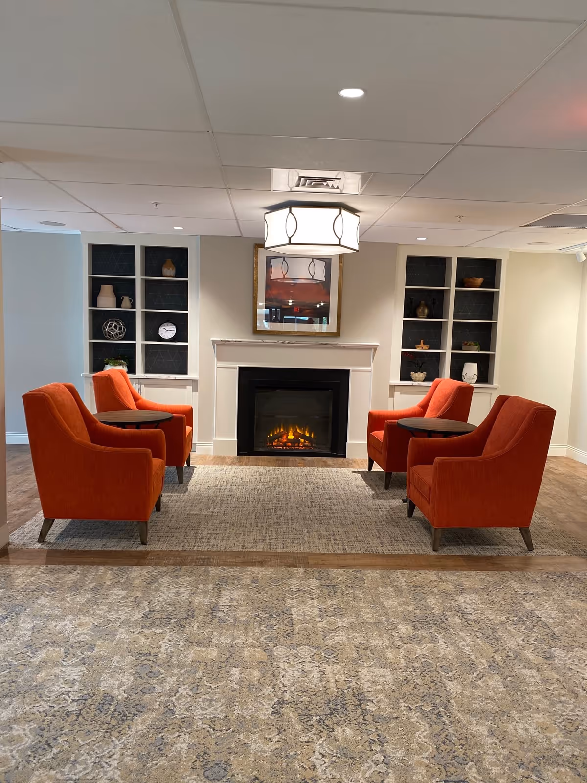 A cozy seating area with four orange armchairs arranged around two small round tables, positioned on a rug in front of a white fireplace with a lit electric fire. Behind the fireplace are built-in shelves with decorative items and a framed picture hanging above the mantel. The ceiling has recessed lighting and a central decorative light fixture.