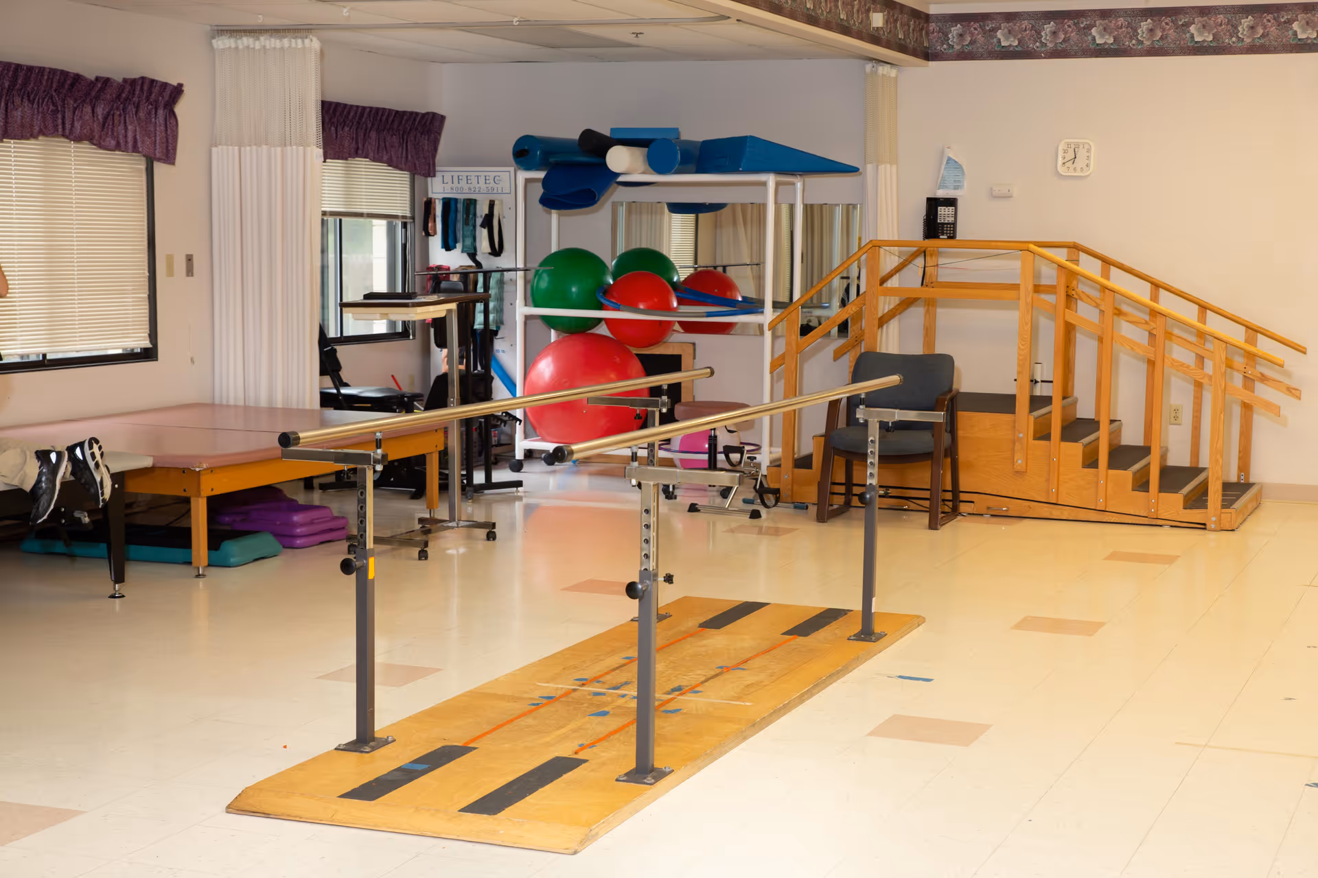 Physical therapy room with parallel bars for walking exercises, a wooden staircase with handrails for step training, exercise balls, therapy tables, and chairs. The room has windows with blinds and purple valances, and light-colored flooring with some colored tiles.