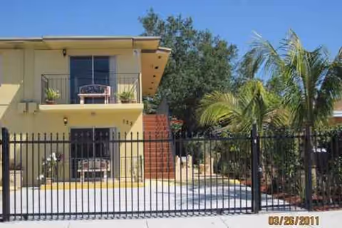 Two-story yellow assisted living facility behind a black metal gate with palm trees nearby.