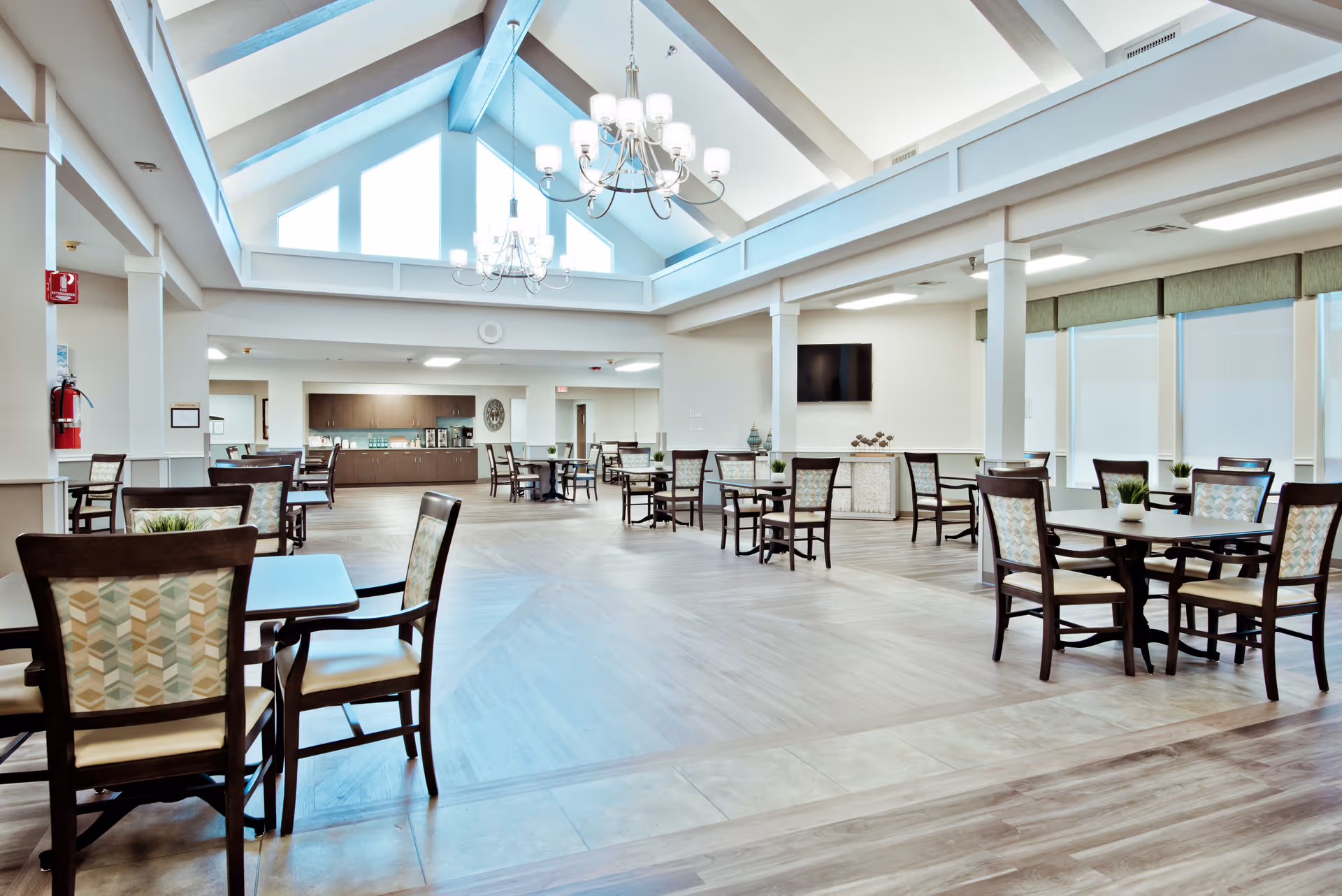 A spacious and well-lit dining area with multiple wooden tables and chairs arranged neatly. The room features large windows with blinds, a high ceiling with exposed beams, and two chandeliers hanging from the ceiling. There is a flat-screen TV mounted on the wall and a counter area in the background with cabinets and a clock.