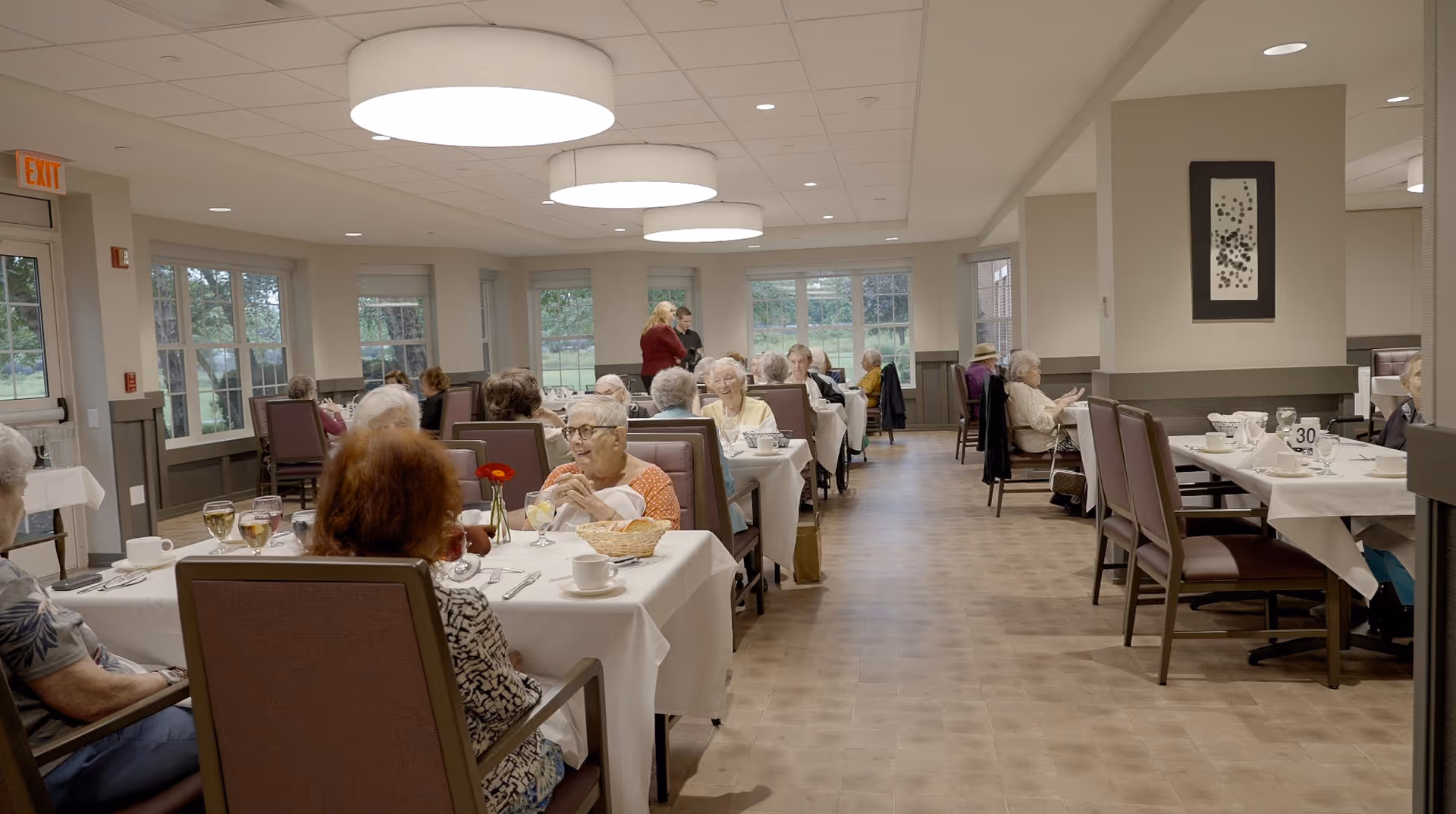A dining room in a senior living facility with elderly residents seated at tables covered with white tablecloths. The room has large windows letting in natural light, and several round ceiling lights. People are engaged in conversation and dining.