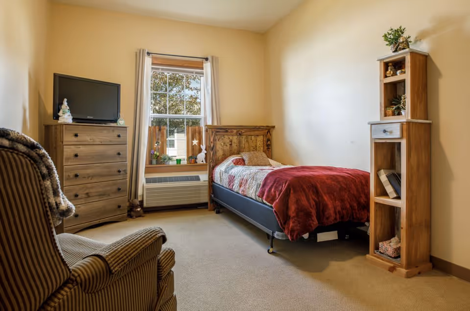 Small furnished bedroom with a single bed, wooden dresser and TV, armchair, window and shelving unit.