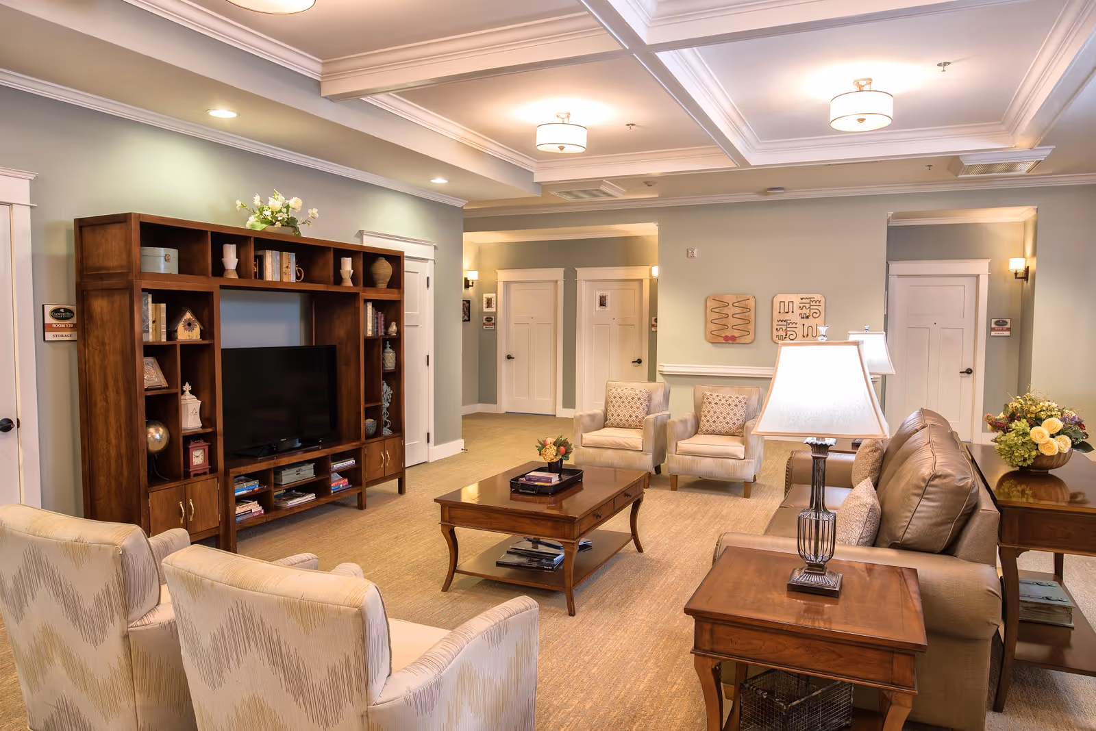 A cozy senior living facility common area with comfortable seating including a leather sofa, two upholstered armchairs, and two patterned chairs. There is a wooden coffee table and side tables with lamps. A large wooden entertainment center with a TV and decorative items is against one wall. The room has soft lighting from ceiling fixtures and wall sconces, with light-colored walls and carpeted flooring.