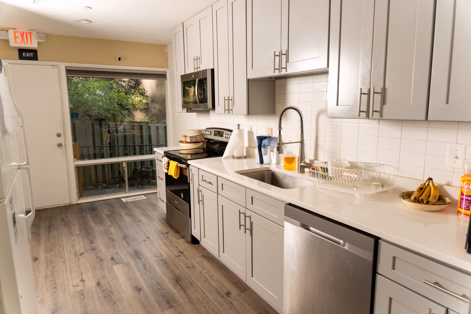 A modern kitchen with light gray cabinets, a stainless steel dishwasher, stove, and microwave. The countertop has a sink with a flexible faucet, a paper towel holder, a water filter pitcher, a dish drying rack, a bowl of bananas, and a bottle of Metamucil. There is a door with an exit sign above it and a large window showing a fenced outdoor area with greenery.