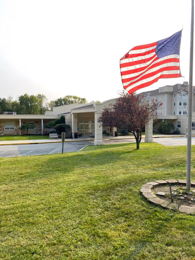 An American flag waves on a flagpole in front of a lawn and the entrance to a multi-story senior living building.