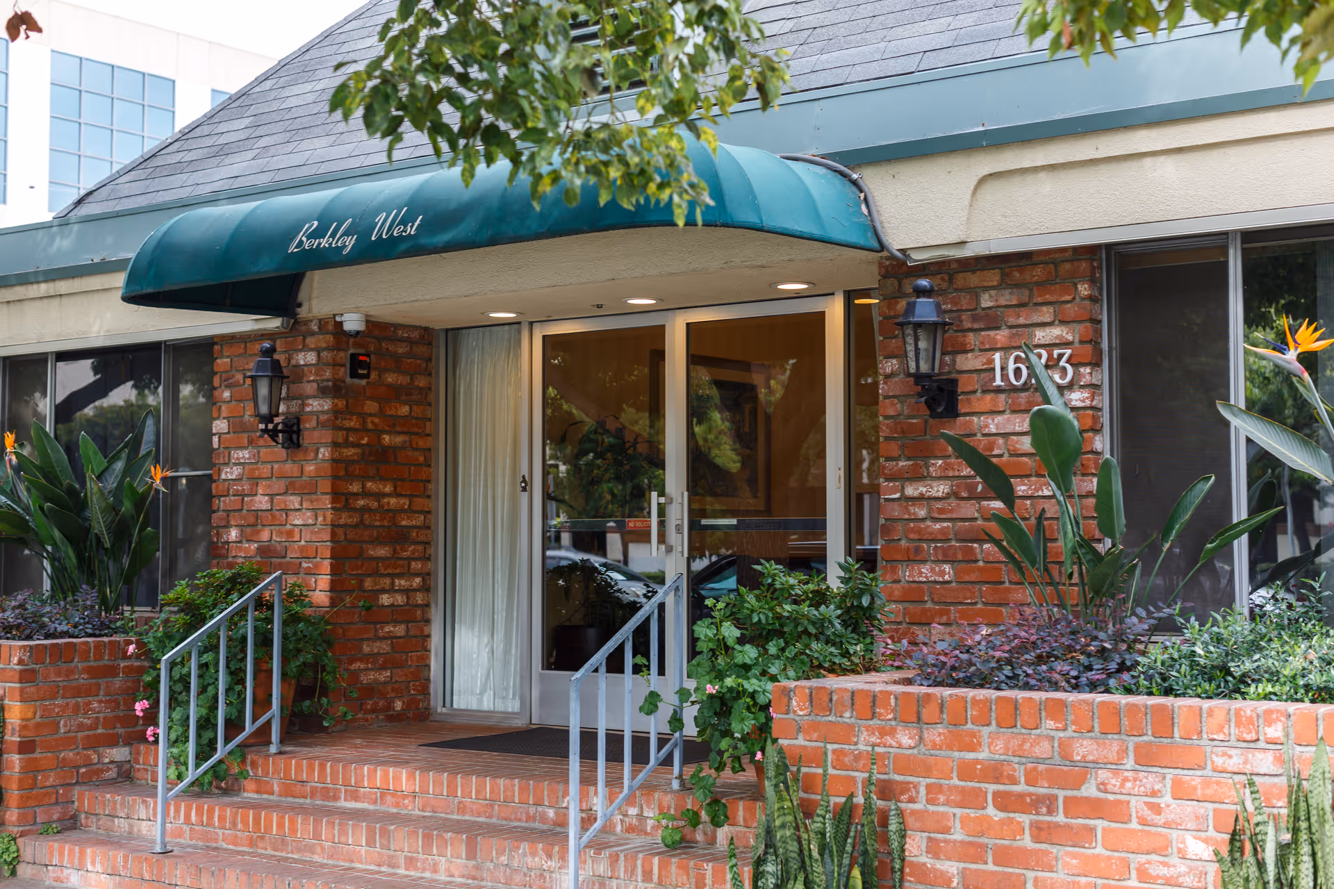 Entrance of Berkley West Healthcare Center showing a brick building with a green awning above the glass double doors. There are plants and shrubs in brick planters on either side of the steps leading up to the entrance. The building number 1623 is visible on the right side.