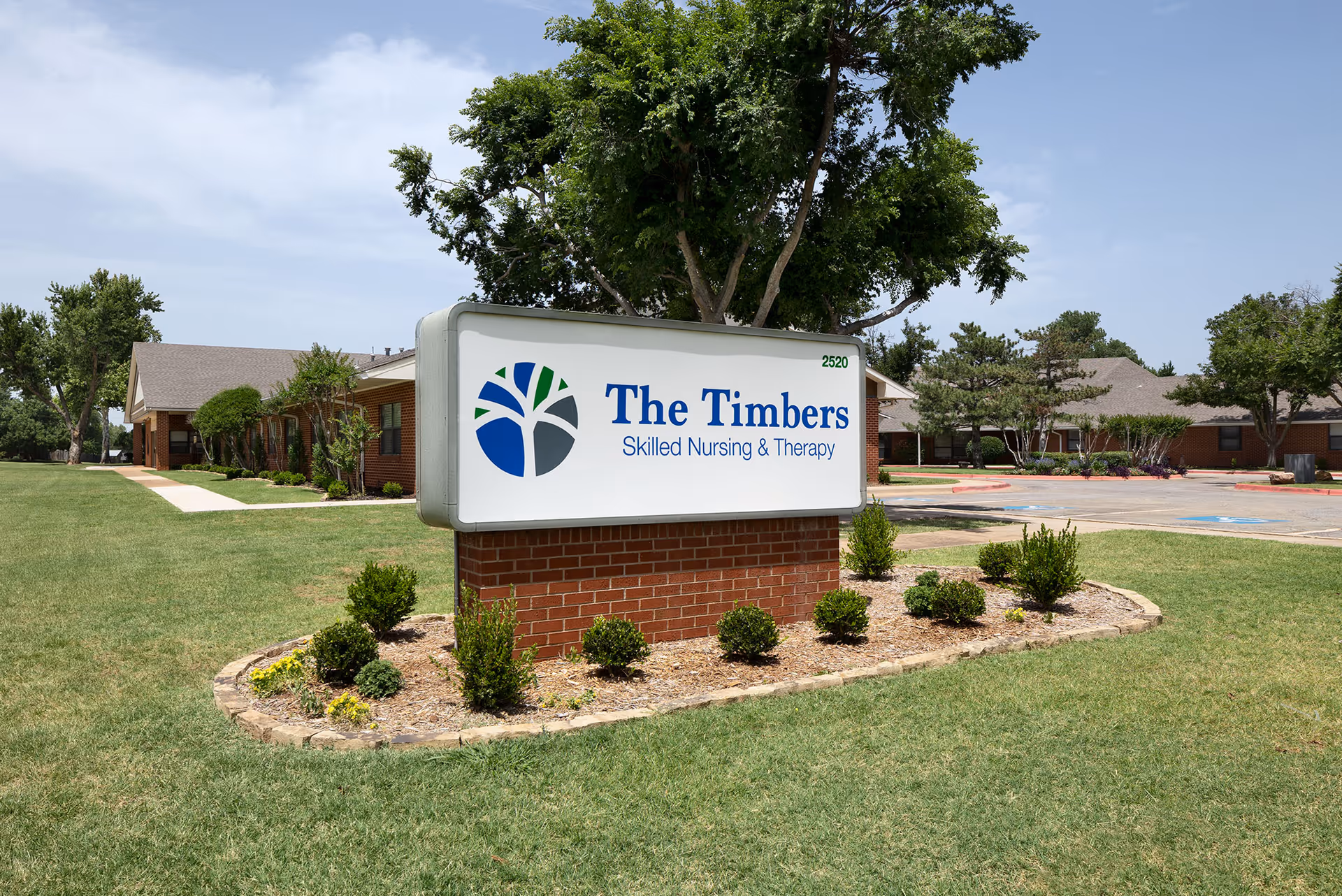 Outdoor view of The Timbers Skilled Nursing & Therapy facility sign on a brick base surrounded by small shrubs and grass, with the building and trees in the background under a partly cloudy sky.