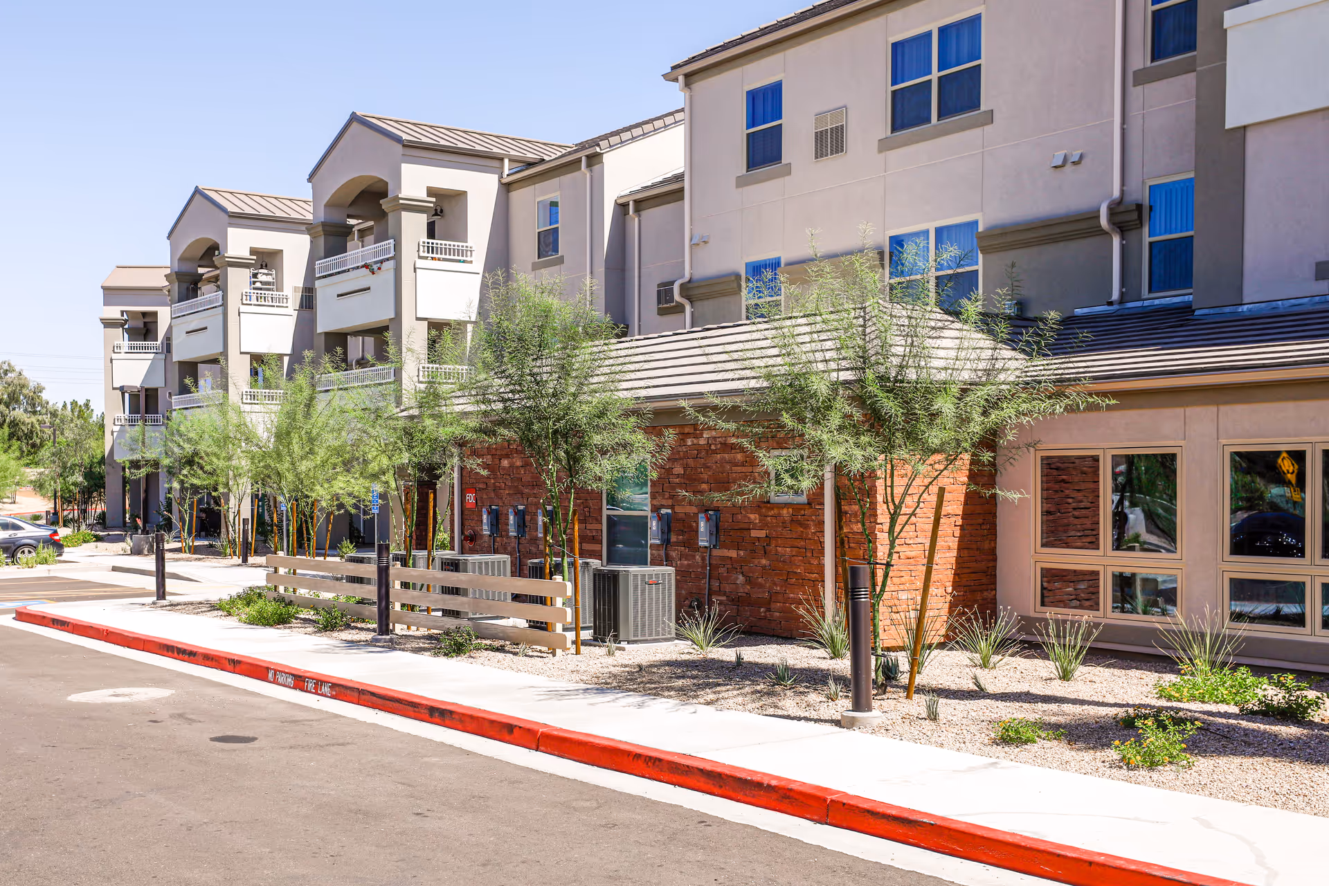 Exterior view of a multi-story senior living facility with balconies, small trees, and landscaping along the sidewalk under a clear blue sky.