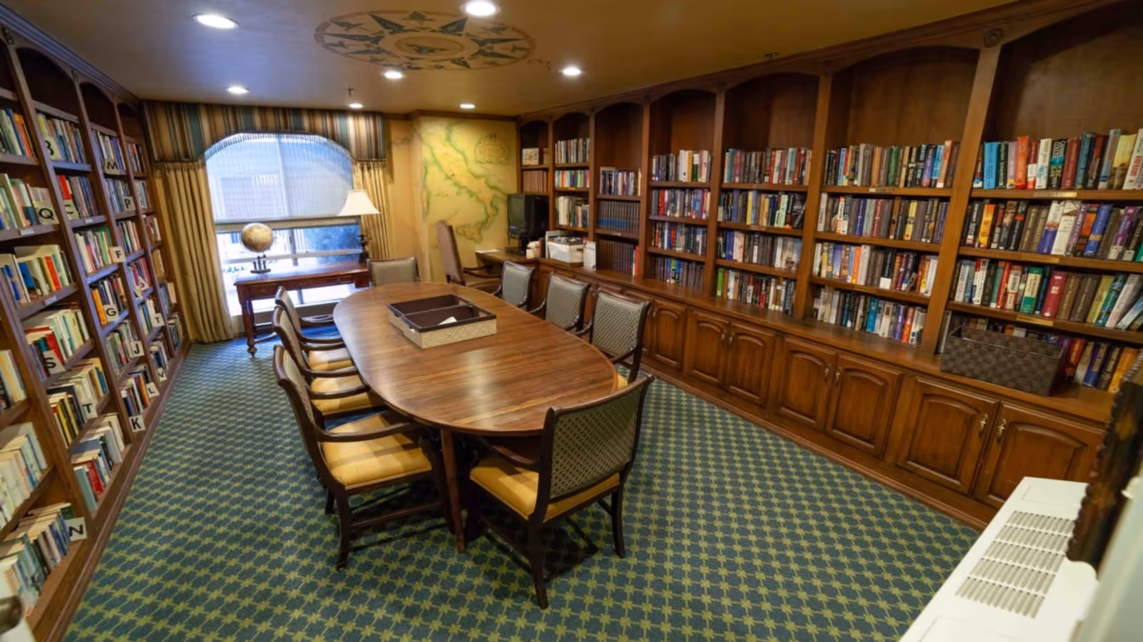 Wood-paneled library/meeting room with a long oval table surrounded by chairs, built-in bookshelves, and patterned carpet.