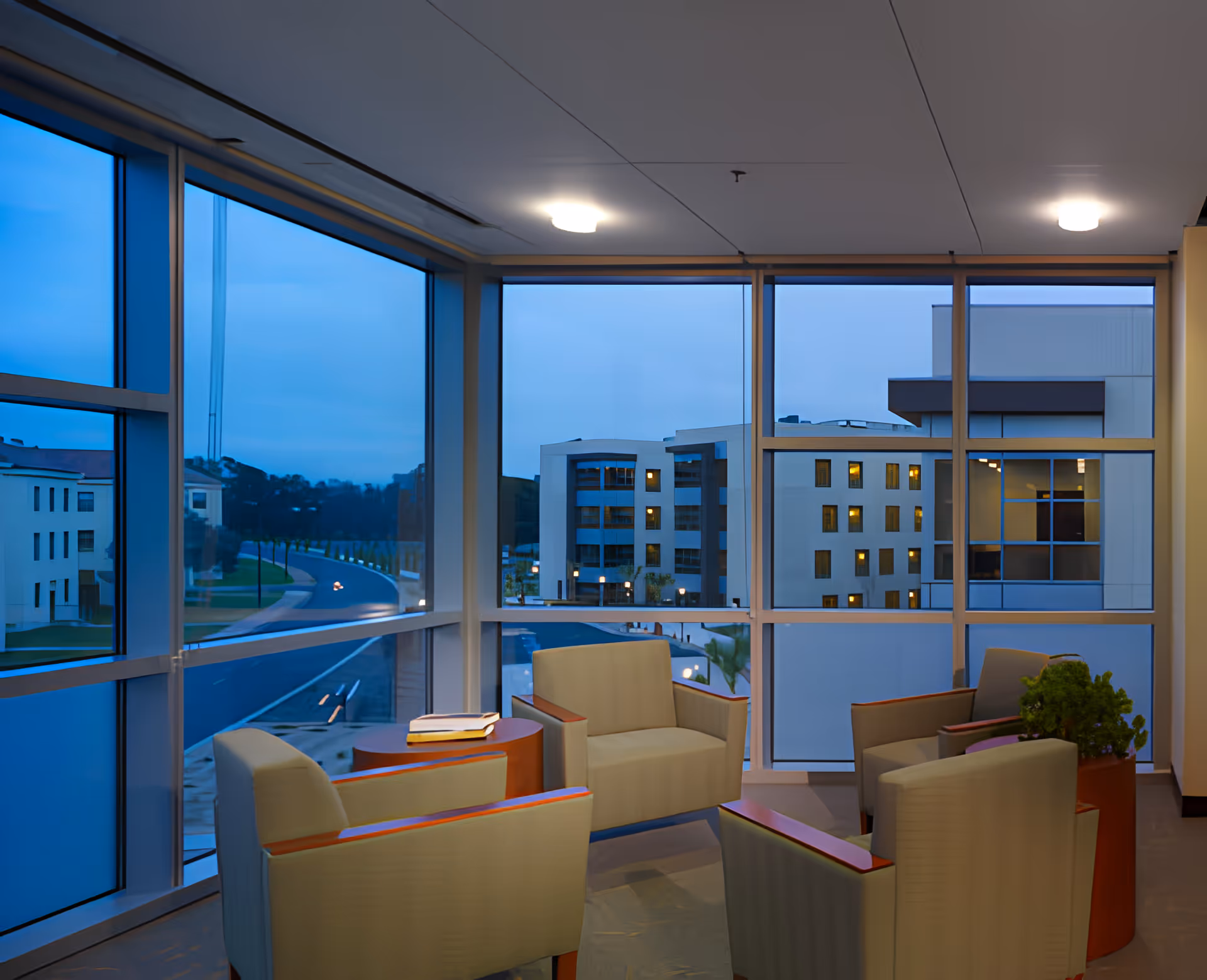 A cozy seating area inside a building with four beige armchairs arranged around a small round wooden table with books on it. Large floor-to-ceiling windows provide a view of other buildings and a road outside during dusk or early evening.