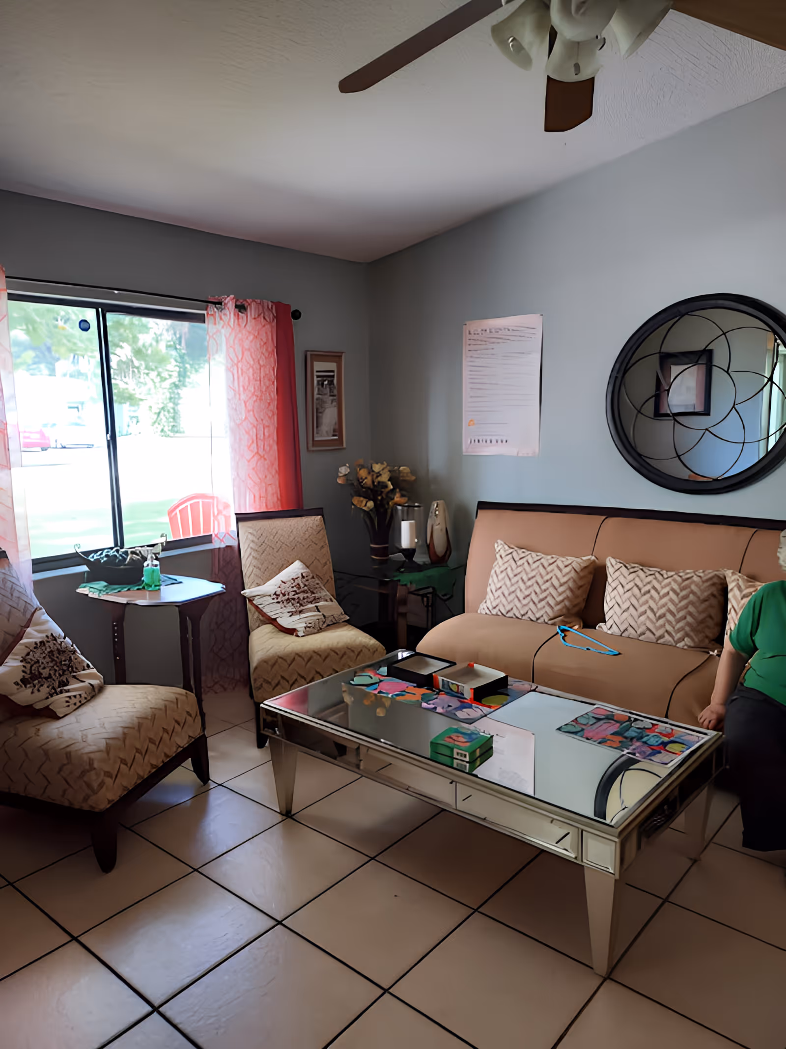 A cozy living room with a beige sofa adorned with three patterned cushions, two matching armchairs with decorative pillows, a mirrored coffee table with colorful items on top, a round decorative wall mirror, and a window with pink curtains letting in natural light. A ceiling fan is visible above, and part of a person wearing a green shirt is seated on the right side of the sofa.