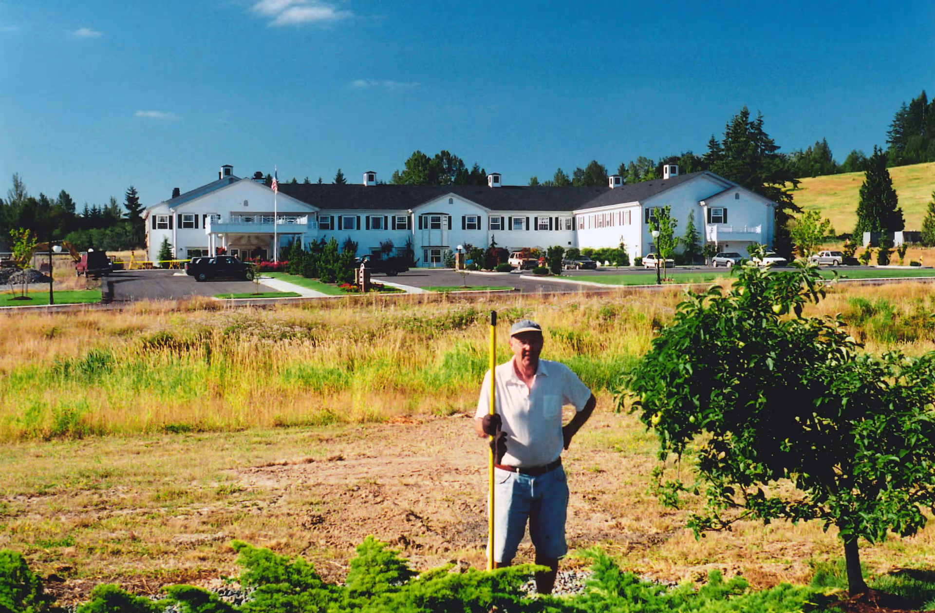 A man holding a long-handled tool stands in a grassy field with a large white senior living building in the background.