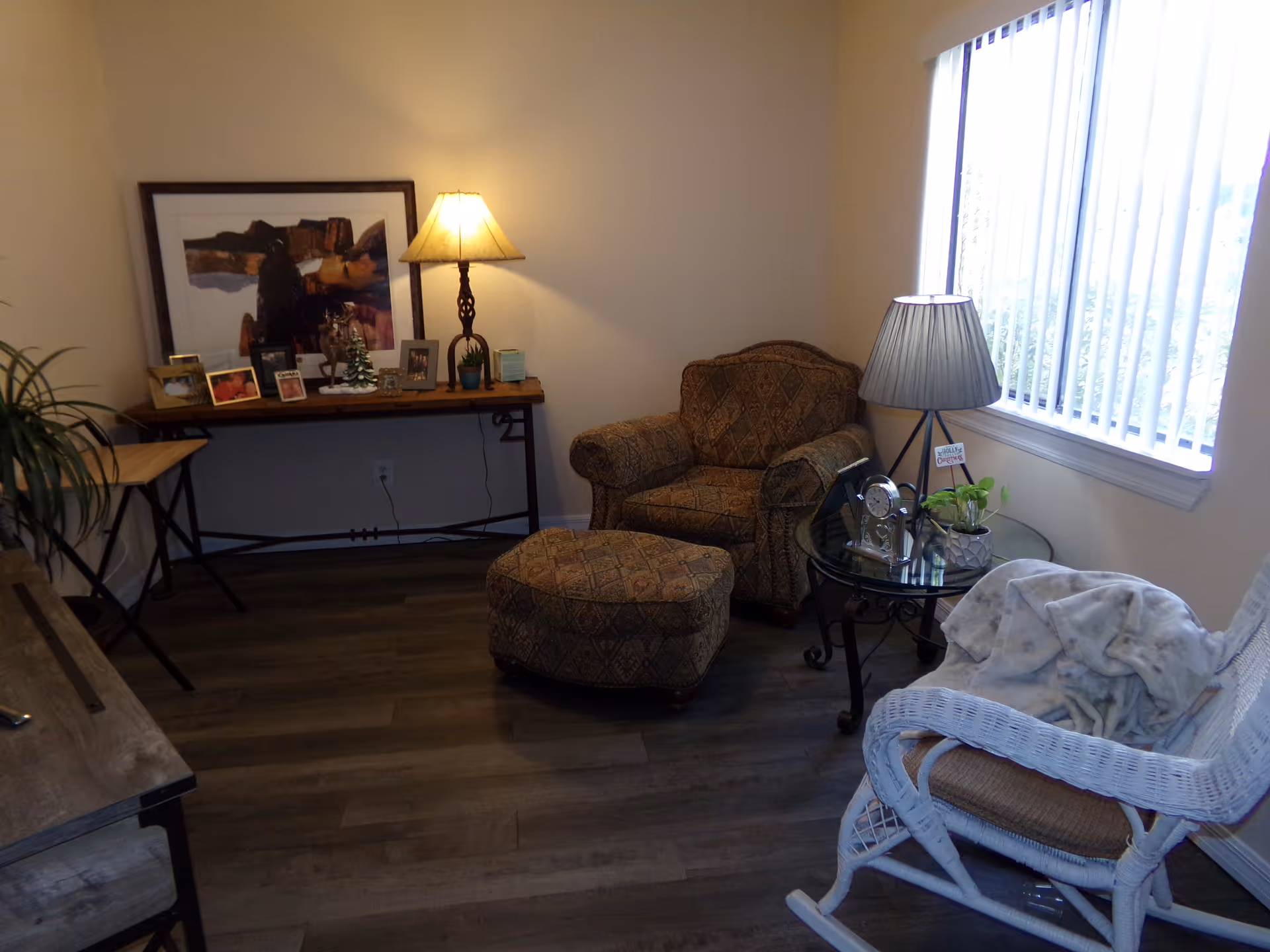 Cozy sitting area with a patterned armchair and ottoman, wicker rocking chair, side table with a lamp, and a console table with framed photos.