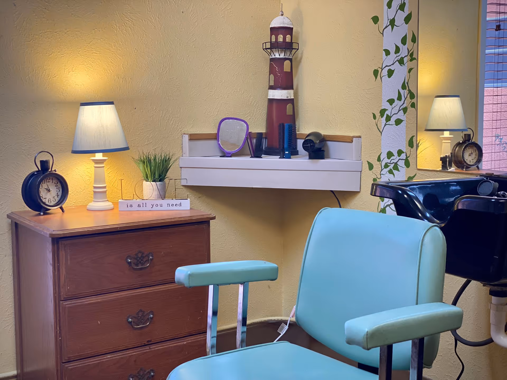 A cozy corner of a room with a vintage wooden dresser topped with a lamp, an old-fashioned alarm clock, a small potted plant, and a decorative sign that reads 'Love is all you need.' Above the dresser is a small white shelf holding a lighthouse decoration, a purple hand mirror, a hairbrush, and a hairdryer. In front is a light blue salon chair, and to the right is a black salon sink. A large mirror on the wall reflects part of the scene, and the walls are painted yellow with a leafy vine decoration on a white column.