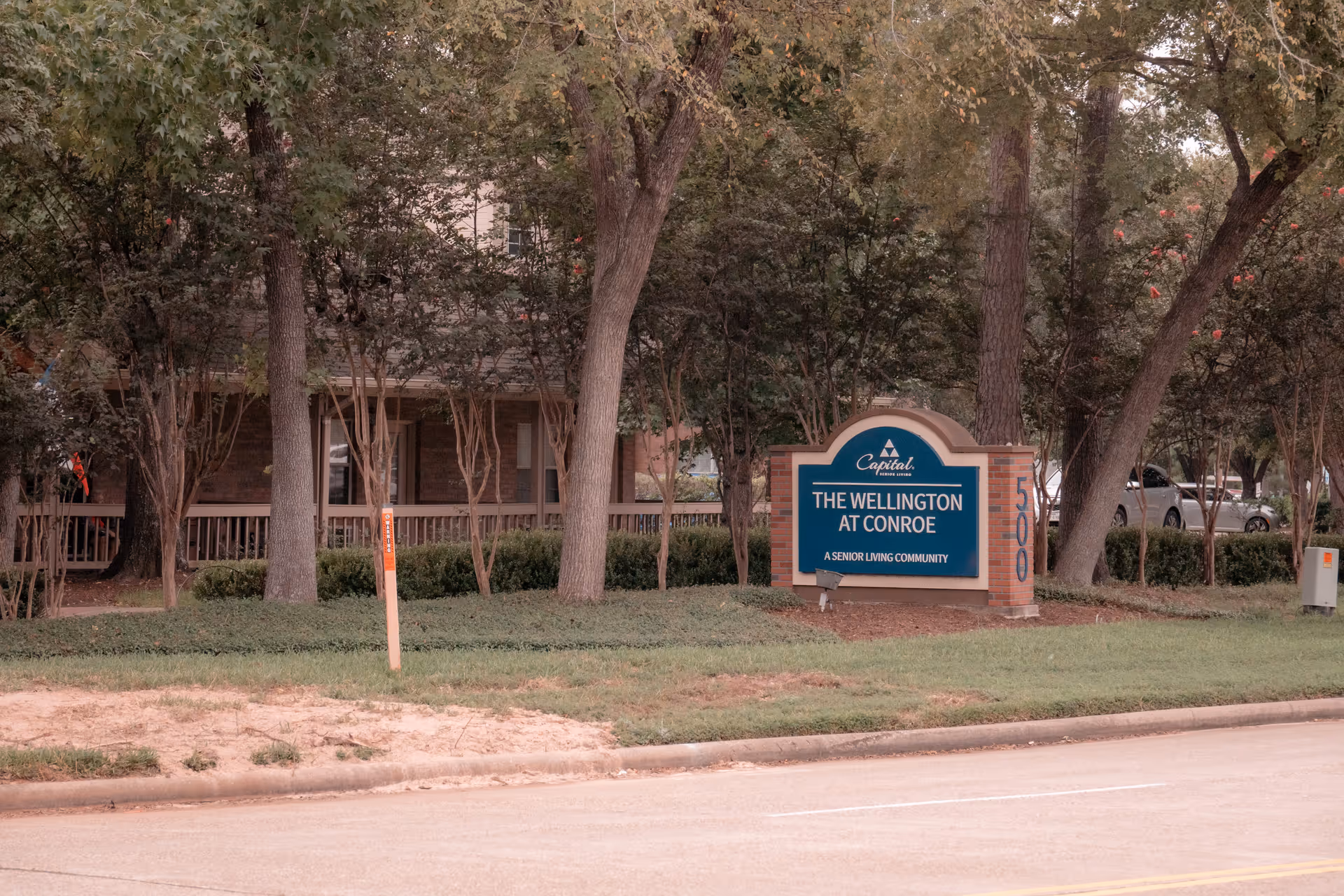 Outdoor view of The Wellington at Conroe senior living community sign surrounded by trees and greenery, with a building and parked cars visible in the background.