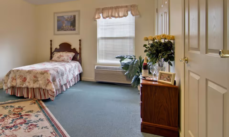 A cozy bedroom with a single bed covered in floral bedding, a wooden headboard, and a matching pillow. There is a window with blinds and a valance letting in natural light. A wooden dresser holds a bouquet of yellow roses, a bottle of wine, two wine glasses, and a framed photo. The room has a green carpet and a floral area rug near the bed. A framed picture hangs on the wall above the bed.