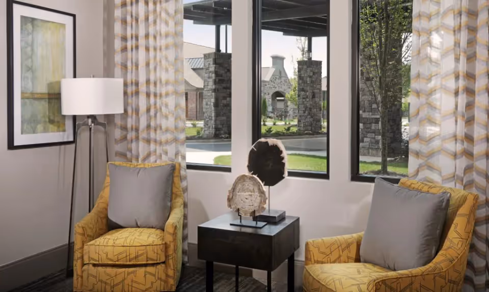 A bright seating area with two yellow patterned armchairs and gray pillows flanking a small black side table and floor lamp in front of windows showing stone pillars outside.