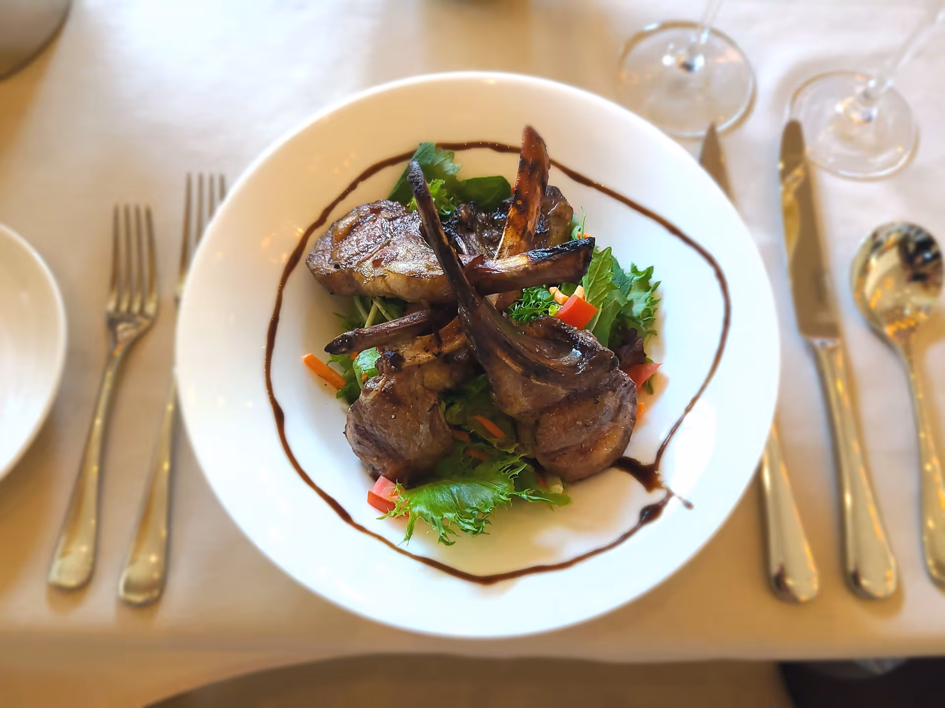 A white plate with grilled lamb chops on mixed greens and a balsamic drizzle, set on a table with utensils and wine glasses.