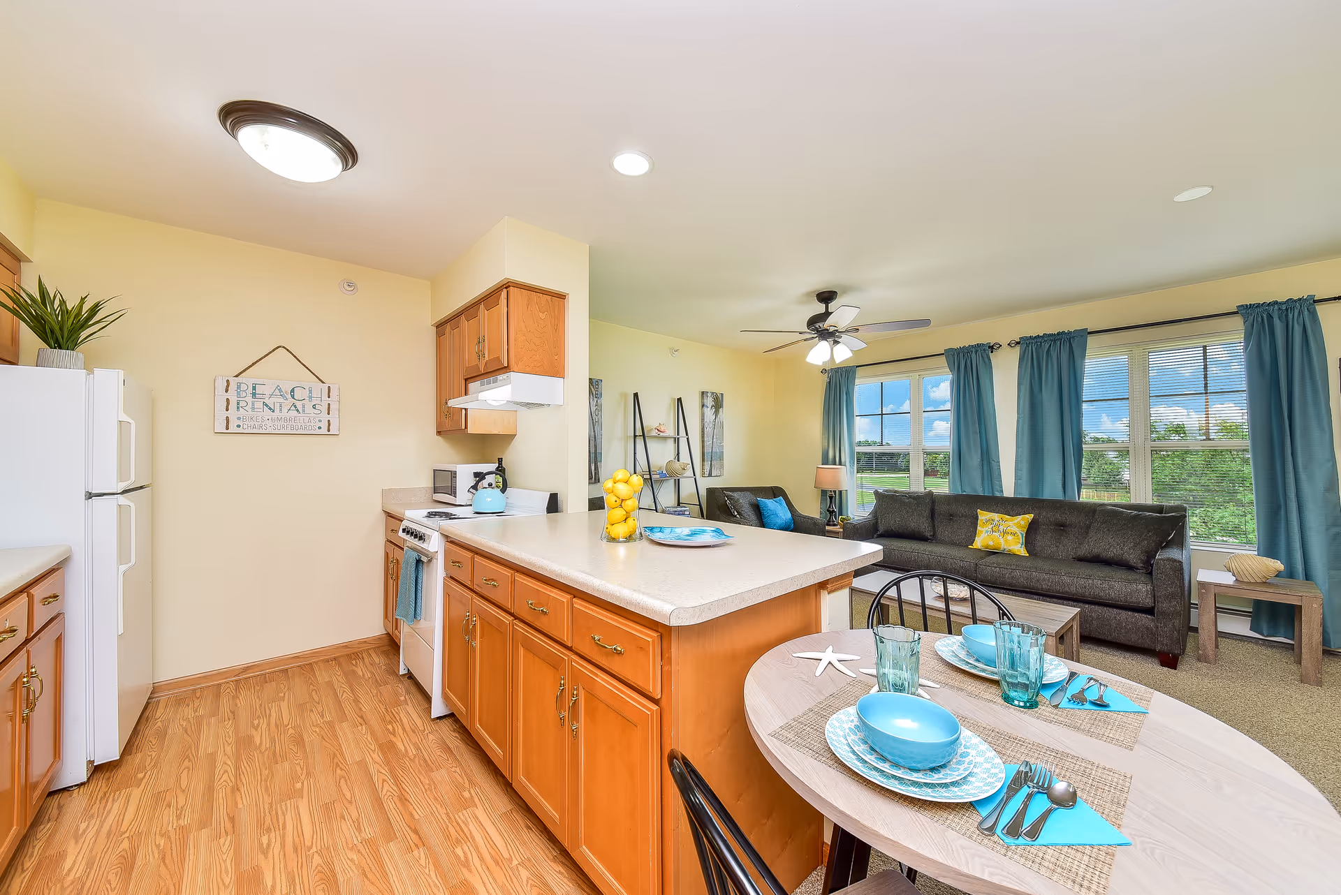 A bright and cozy open-concept living space featuring a kitchen with wooden cabinets, a white refrigerator, stove, and a countertop island. Adjacent to the kitchen is a small dining table set with turquoise dishes and glasses. The living room area has a dark gray sofa with colorful pillows, a ceiling fan with lights, large windows with blue curtains, and a small side table with a lamp. The walls are painted light yellow, and the floor is a mix of wood and carpet.