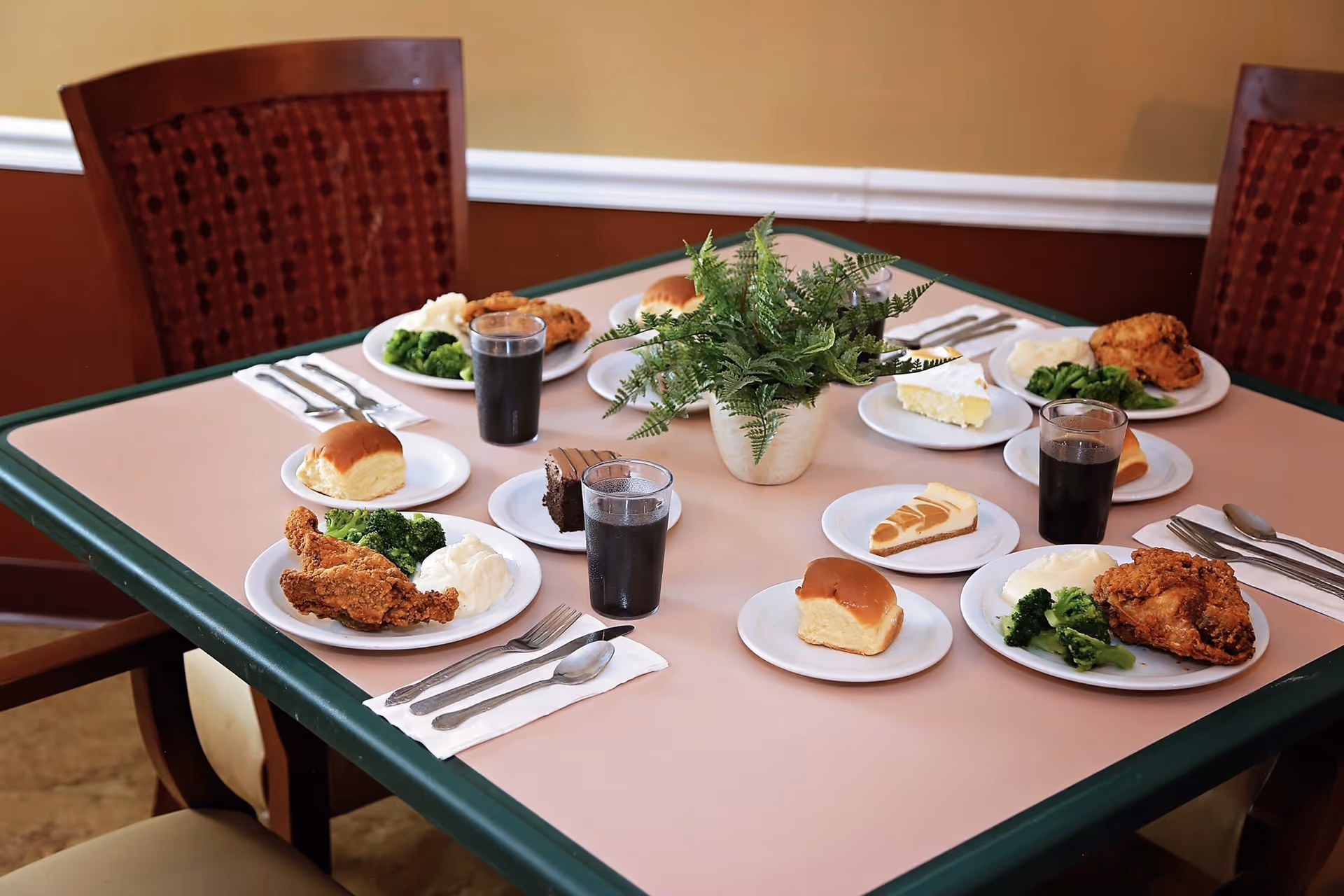 A dining table set with four meals, each including fried chicken, broccoli, mashed potatoes, a bread roll, a slice of dessert, and a glass of dark soda. A green potted plant is placed in the center of the table. The table is surrounded by wooden chairs with red cushioned backs.