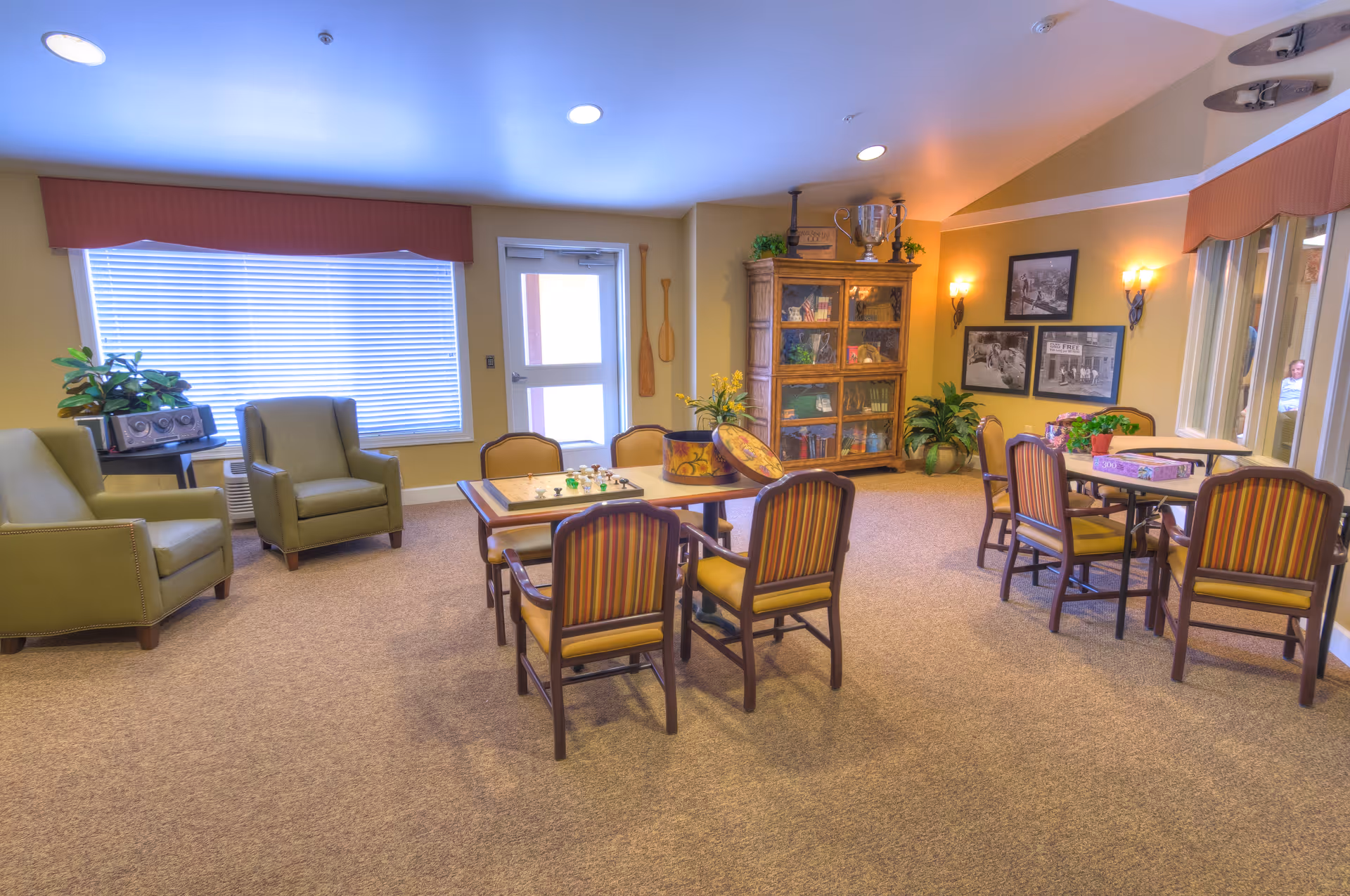 A cozy common area in a memory care facility with several tables and chairs arranged for games and social activities. There are two green armchairs near a window with blinds, a wooden cabinet filled with books and trophies, framed black and white photos on the wall, and some potted plants adding greenery to the space.