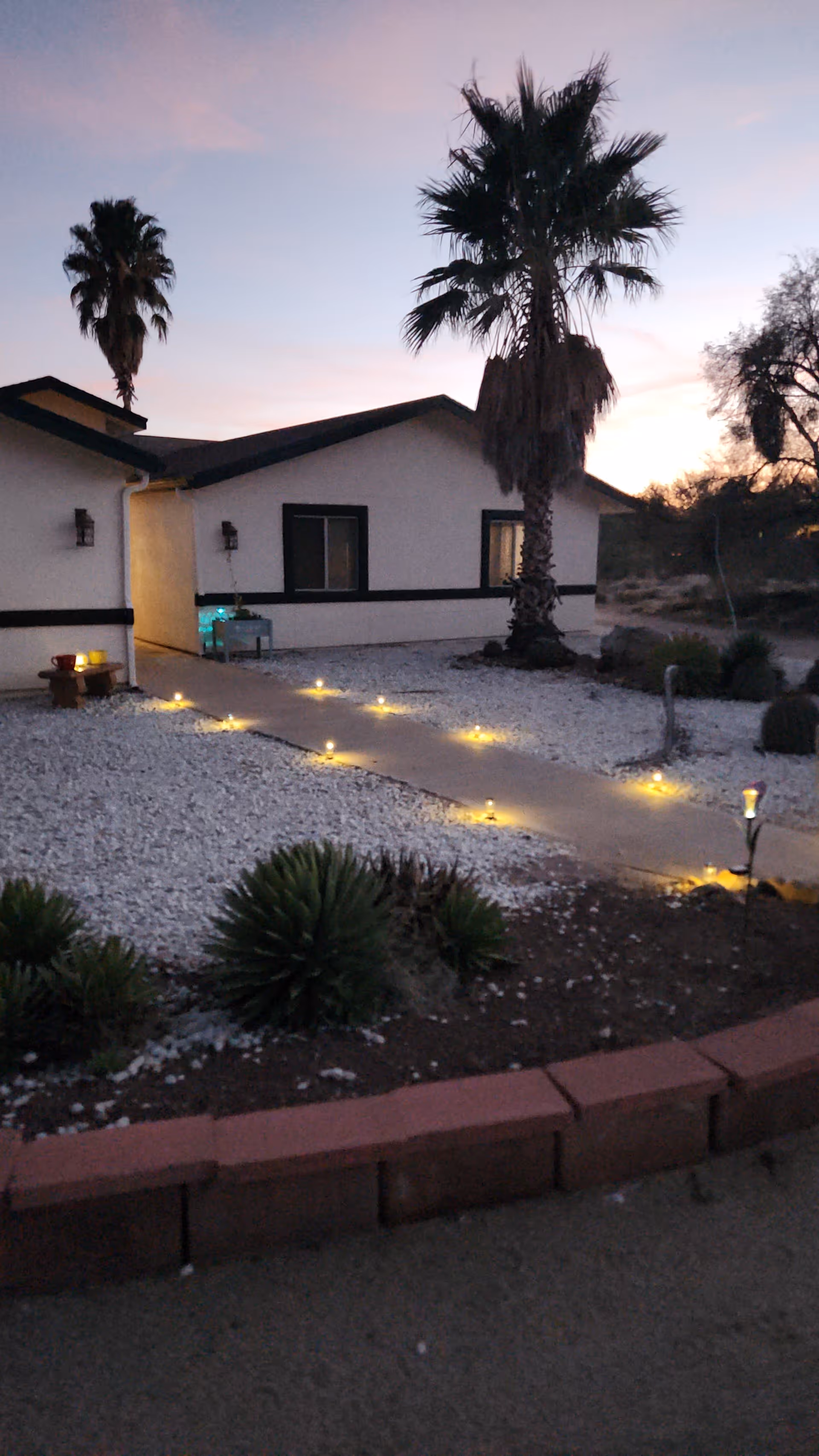 Exterior view of a single-story building at dusk with a pathway lined with small lights leading to the entrance. The building is surrounded by desert landscaping including palm trees, small shrubs, and white gravel. The sky is a gradient of soft colors from the setting sun.