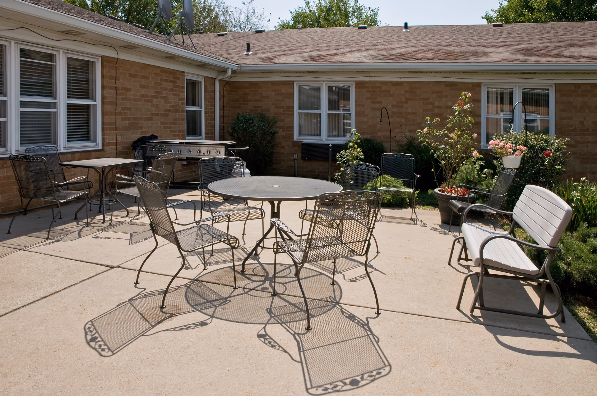 Outdoor patio area with metal tables and chairs arranged on a concrete surface. There is a bench on the right side and a grill against the brick building wall. Various potted plants and flowers are placed near the building windows under a clear sky.