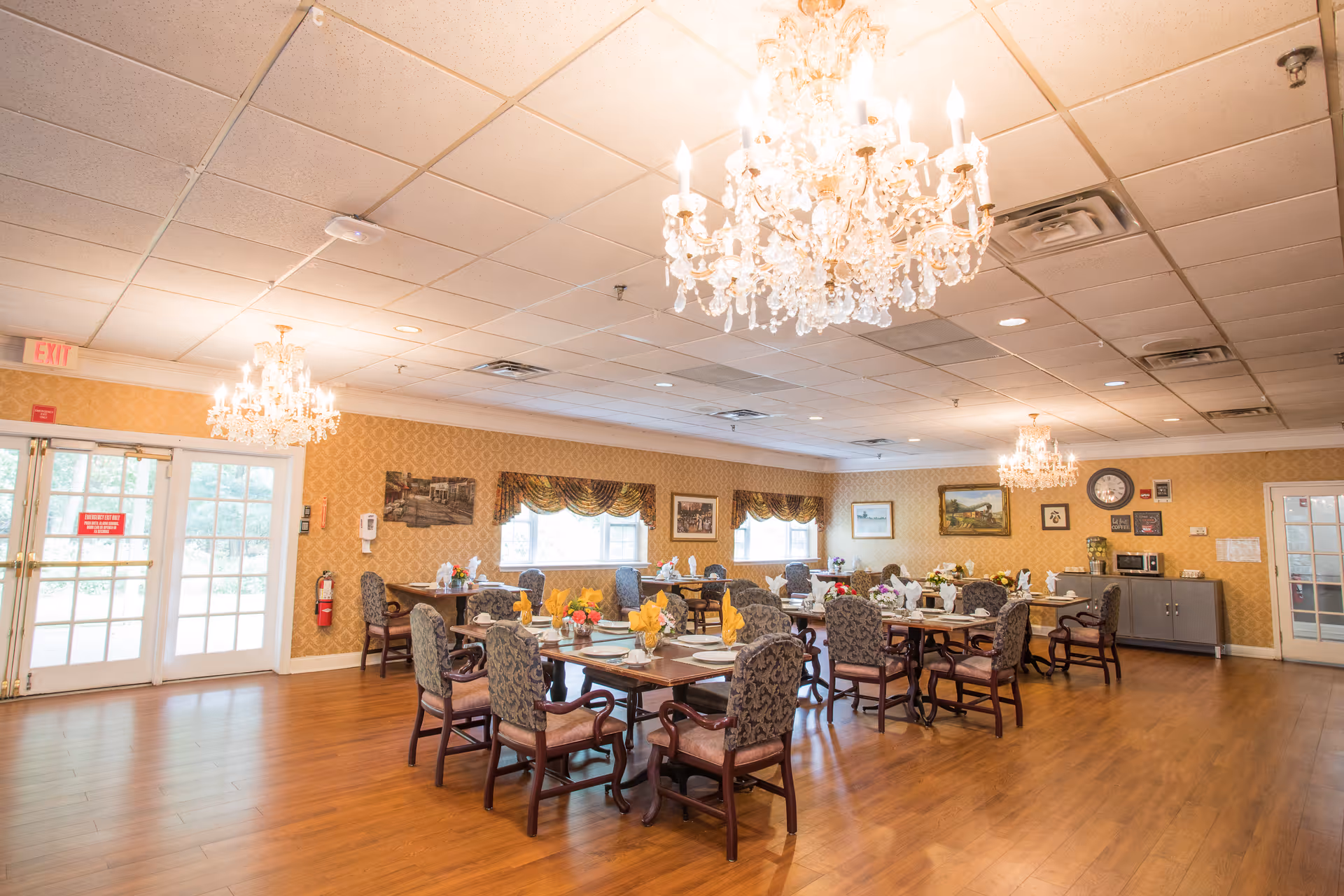 A spacious dining room with multiple tables set with plates, glasses, and yellow napkins. The room features elegant chandeliers, patterned wallpaper, framed artwork on the walls, and large windows with decorative valances. The floor is wooden, and there is a cabinet with a microwave and clock on the far wall. Double glass doors lead outside.
