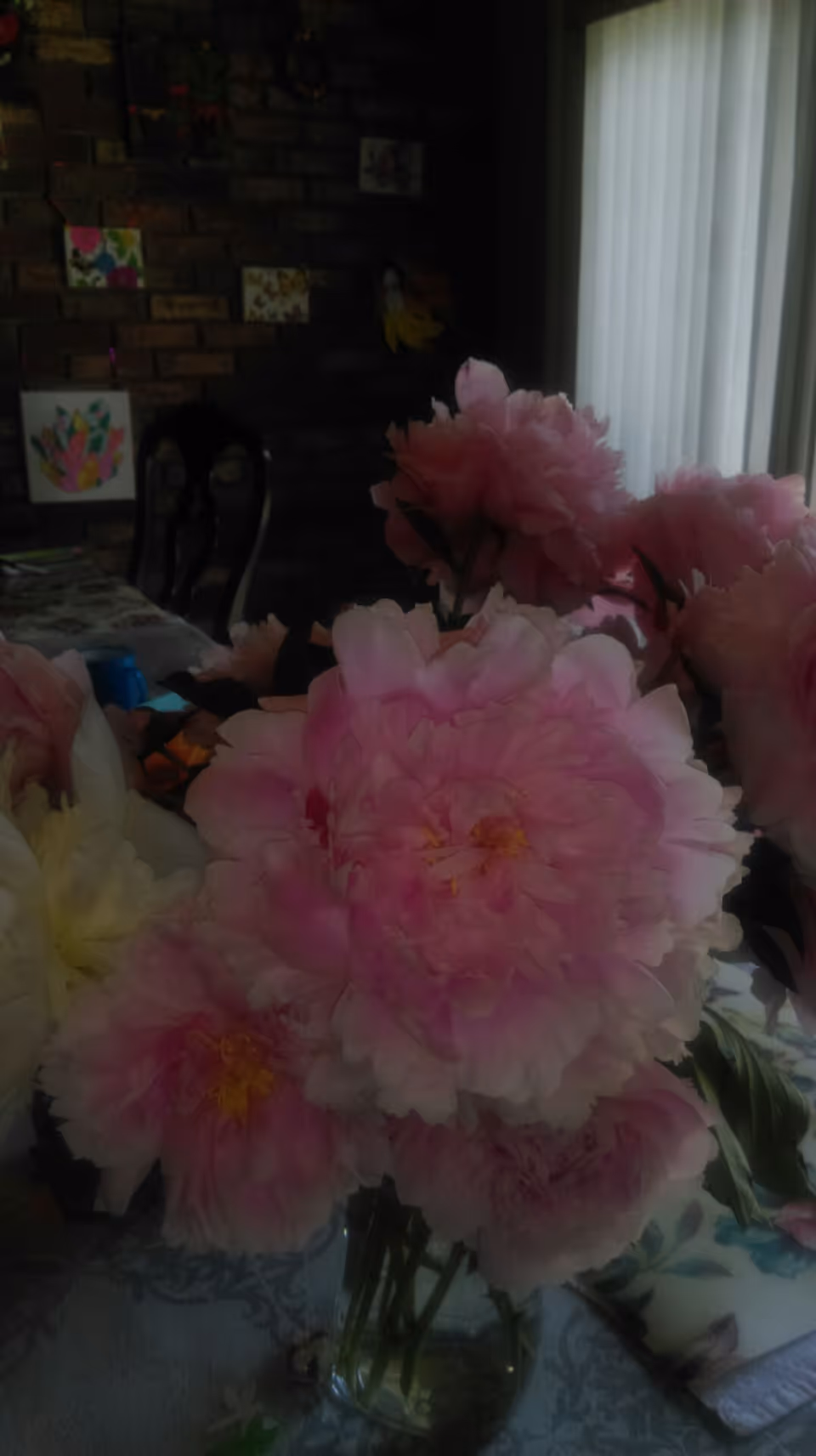 A bouquet of pink peonies in a glass vase on a table inside a room with a brick wall and vertical blinds.
