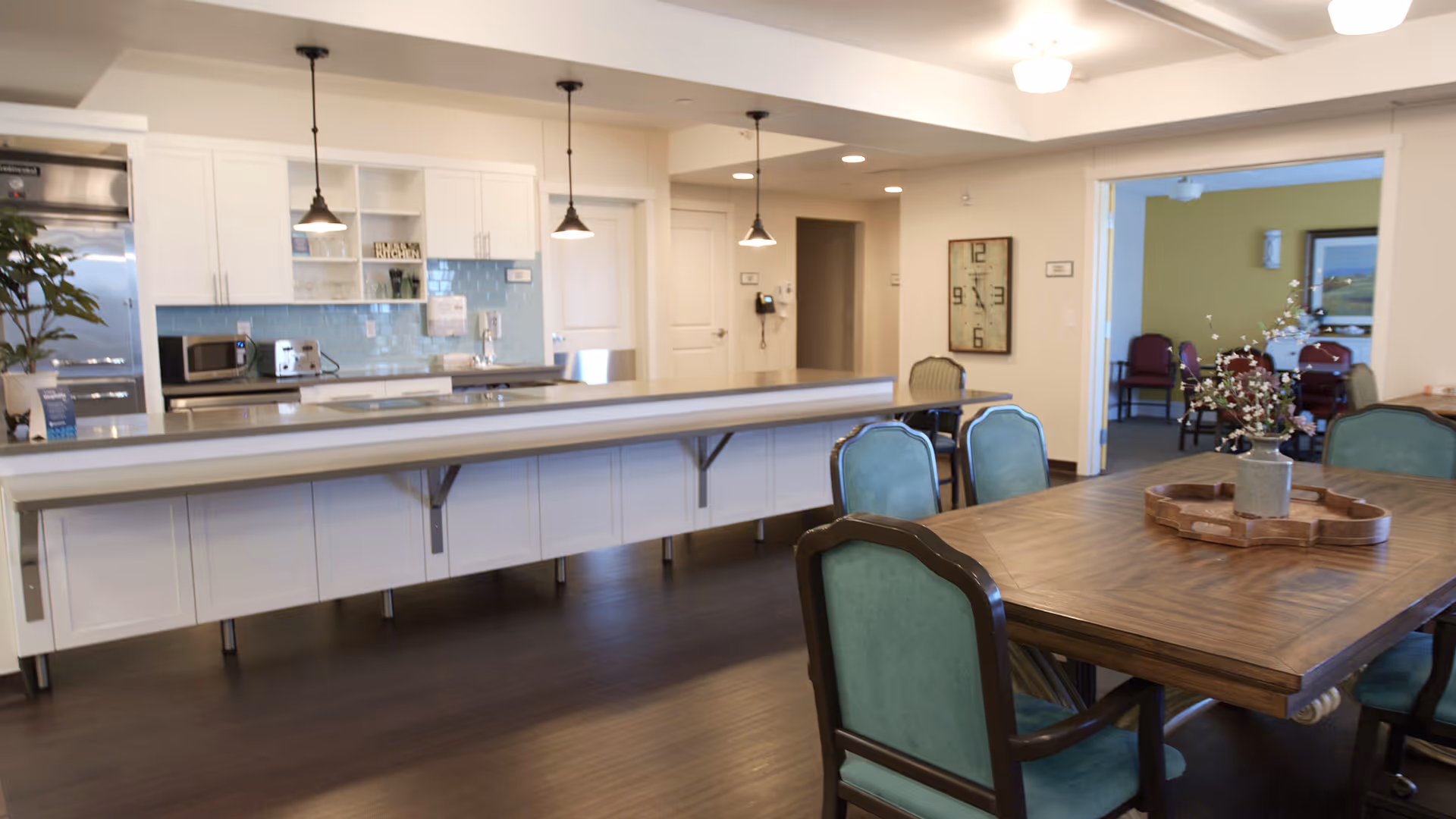 A spacious kitchen and dining area in a senior living community. The kitchen features white cabinets, a light blue tiled backsplash, stainless steel appliances, and pendant lighting. In the foreground, there is a wooden dining table with green upholstered chairs and a decorative centerpiece. In the background, another room with additional seating is visible.