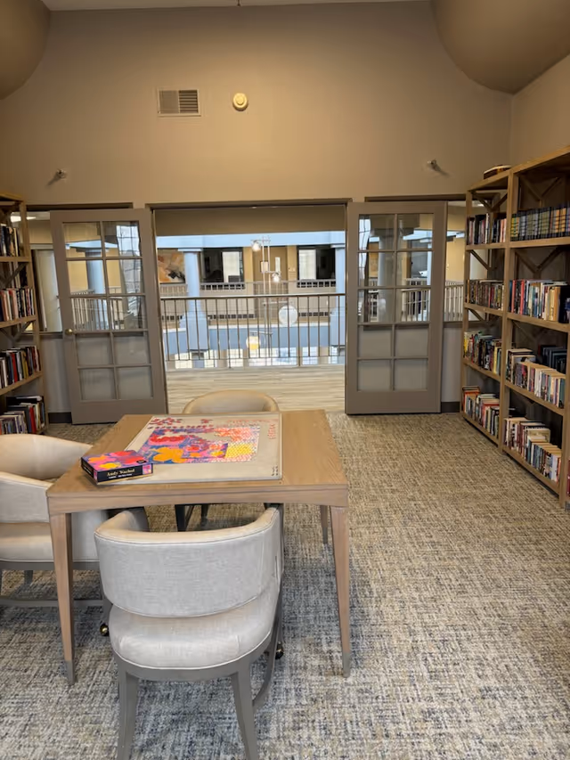 Communal reading area with a table and chairs, bookshelves, and a view of an interior atrium through glass doors.