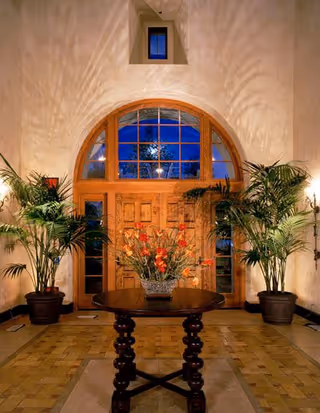 Interior view of an elegant entryway featuring a large wooden arched door with glass panels. Two tall potted plants flank the door, and a round wooden table with a floral arrangement is centered in front of it. Warm lighting casts decorative shadows on the walls.