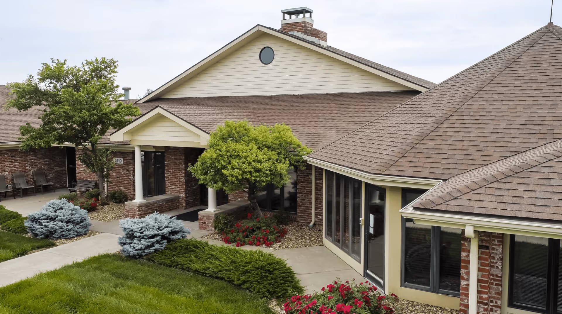 Exterior view of Rolling Hills Assisted Living facility showing a brick building with beige siding, a chimney, multiple windows, a covered entrance with white columns, and well-maintained landscaping including green bushes, small trees, and red flowers.