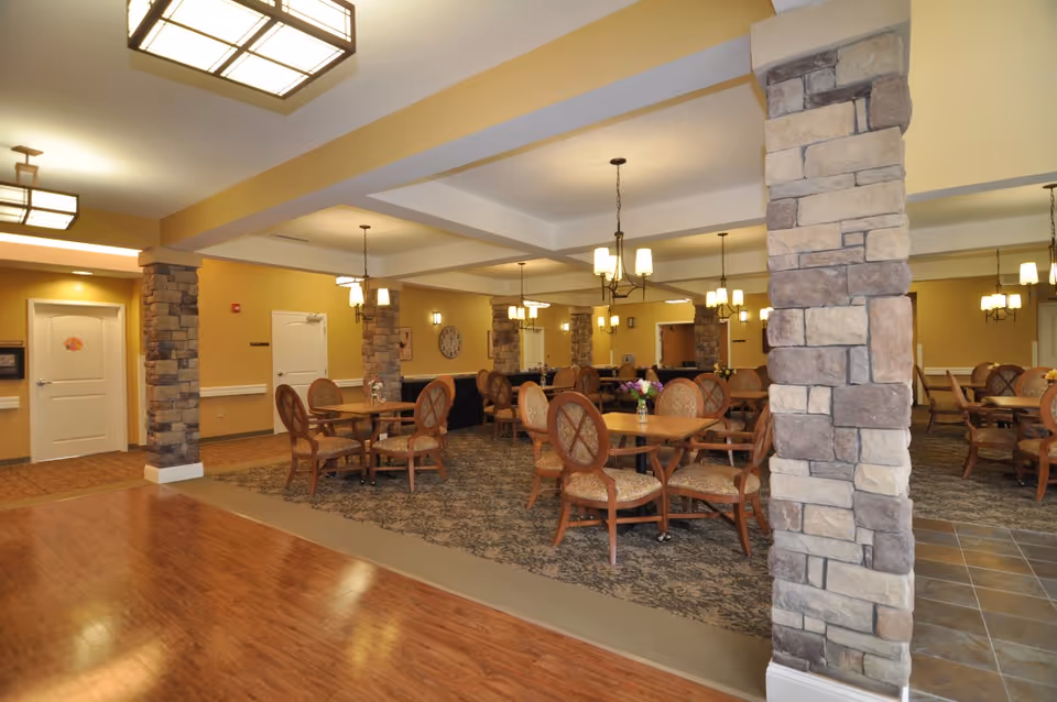 Spacious dining room with round wooden tables and upholstered chairs, stone columns, and warm overhead lighting.