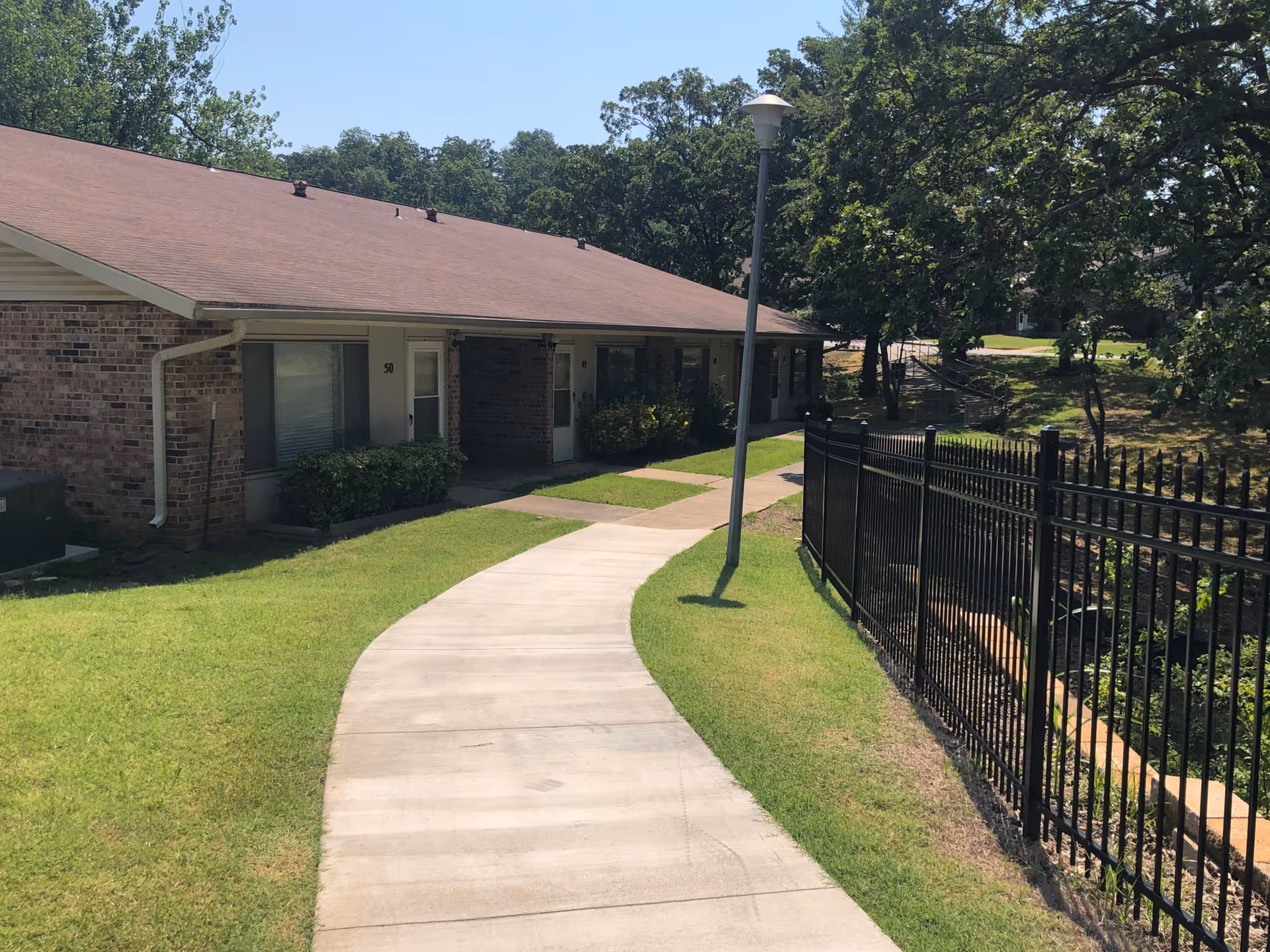 A curved concrete walkway leads past a single-story brick building with a brown roof. The building has several doors and windows, with bushes planted near the walls. On the right side of the walkway, there is a black metal fence and a lamp post. Trees and grass surround the area under a clear blue sky.