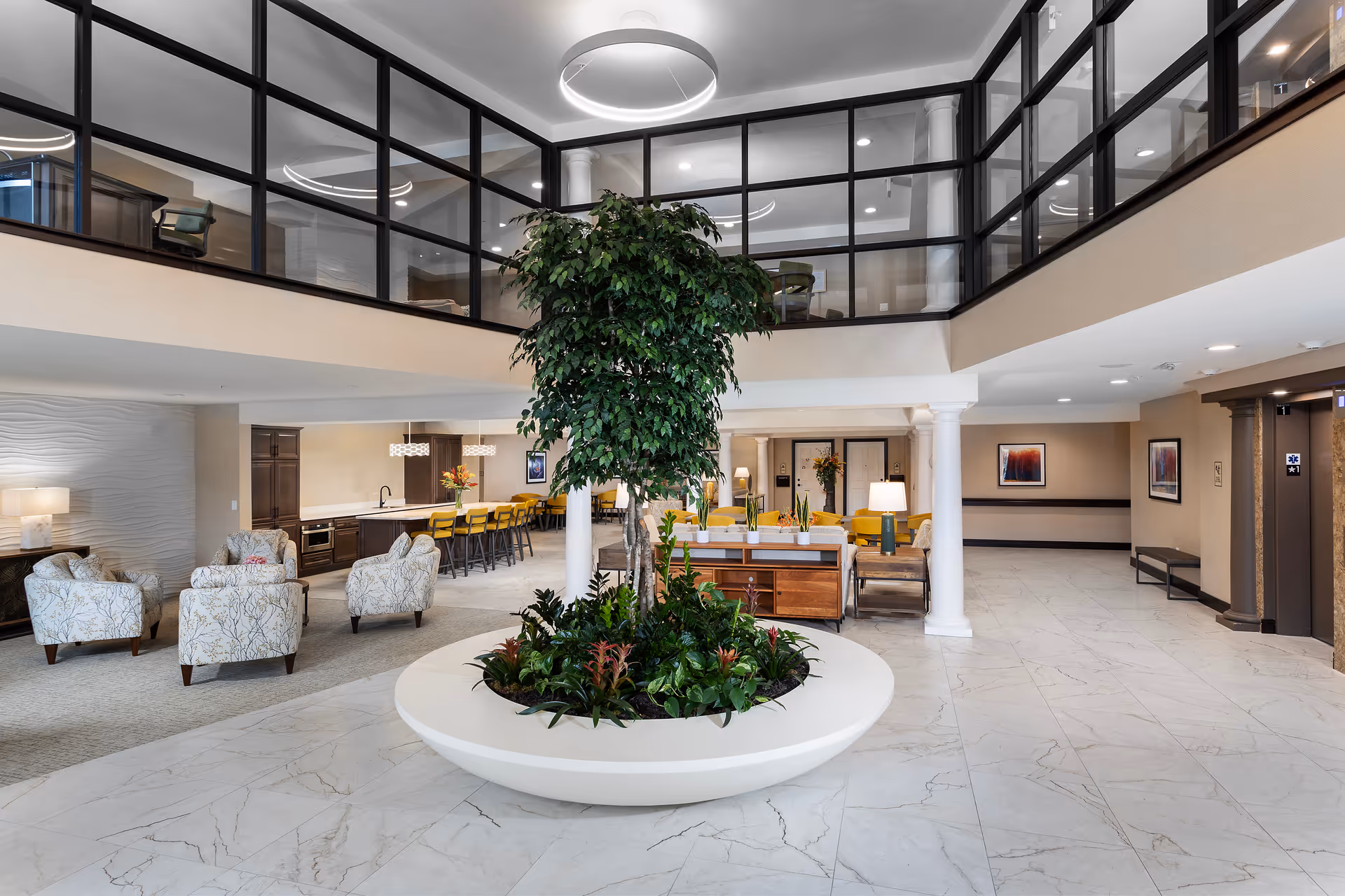 Bright two-story senior living lobby with a large central planter tree, seating areas, and a dining bar.