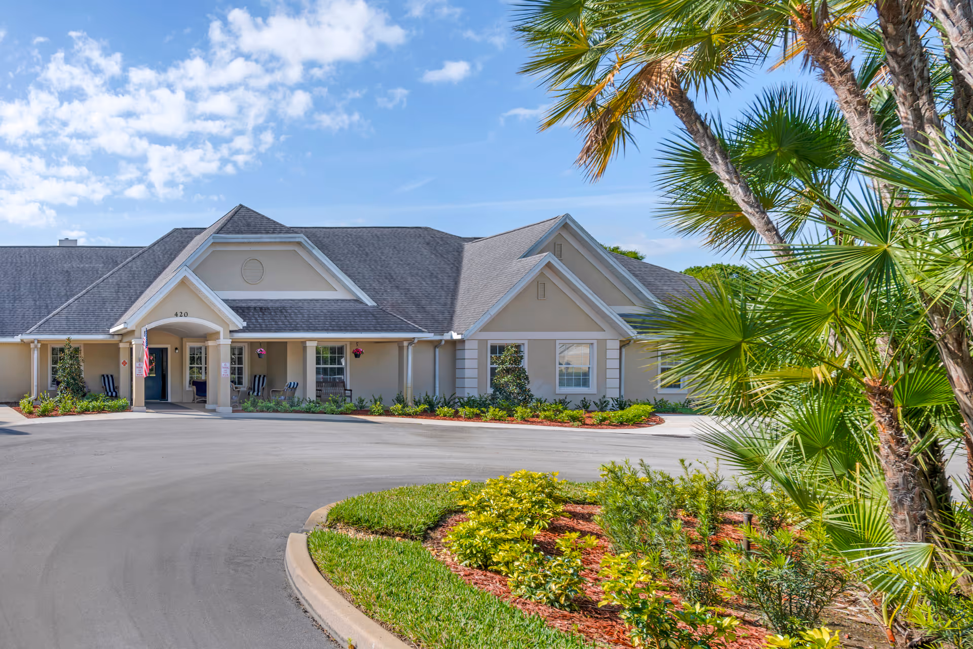 Single-story senior living facility front entrance with a circular driveway, landscaped beds and palm trees under a blue sky.