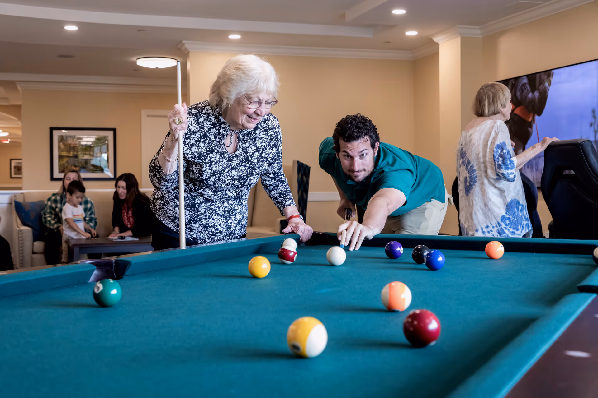 An elderly woman and a younger man playing pool together in a well-lit room. The woman is holding a pool cue and smiling, while the man is aiming to take a shot. In the background, there are other people sitting on a couch and a large TV screen on the wall.