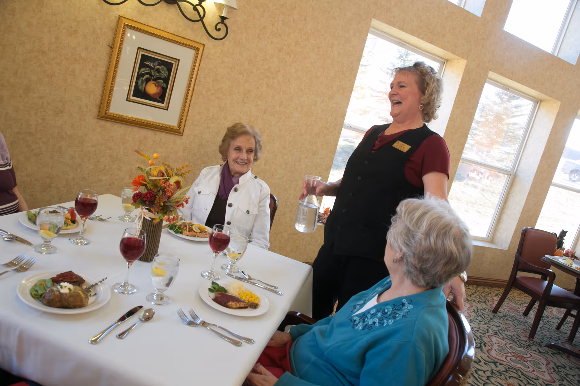 Two elderly women seated at a dining table with plates of food and glasses of water and red beverage, smiling and interacting with a standing female server holding a water pitcher in a well-lit dining room with large windows and framed artwork on the wall.