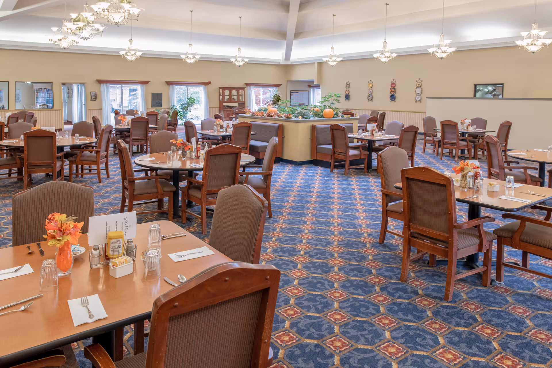 Spacious dining room with multiple wooden tables and chairs arranged neatly. Tables are set with glasses, napkins, utensils, and small floral centerpieces. The room features patterned blue carpet, large windows letting in natural light, and decorative ceiling lights. There is a central divider with plants and seasonal decorations.