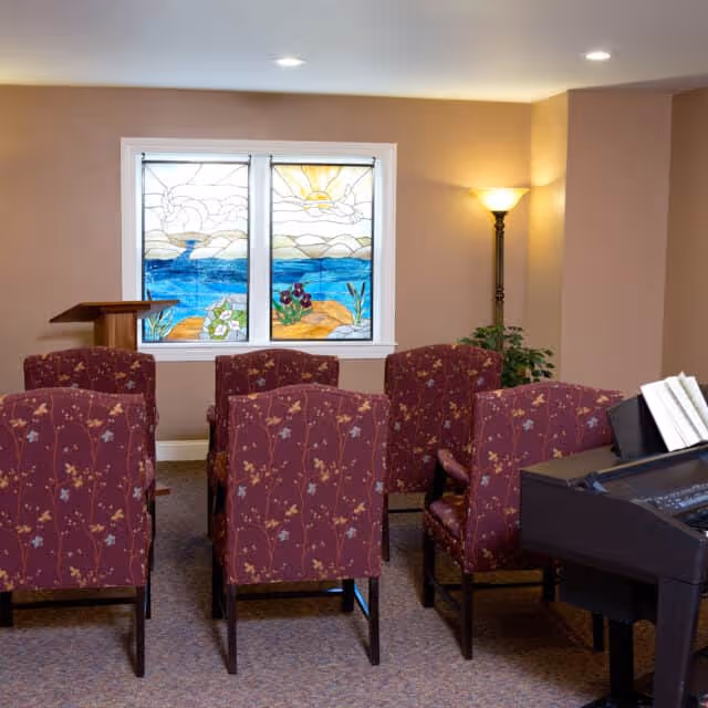 A small room with six upholstered chairs arranged in two rows facing a wooden podium. Behind the podium are two stained glass windows depicting a scenic view with a sun, clouds, water, and flowers. To the right, there is a floor lamp and a piano with sheet music on it. The walls are painted beige.