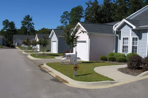 A row of single-story residential buildings with attached garages along a paved street. Each unit has a mailbox and small landscaped front yard with bushes and trees. The sky is clear and there are tall trees in the background.